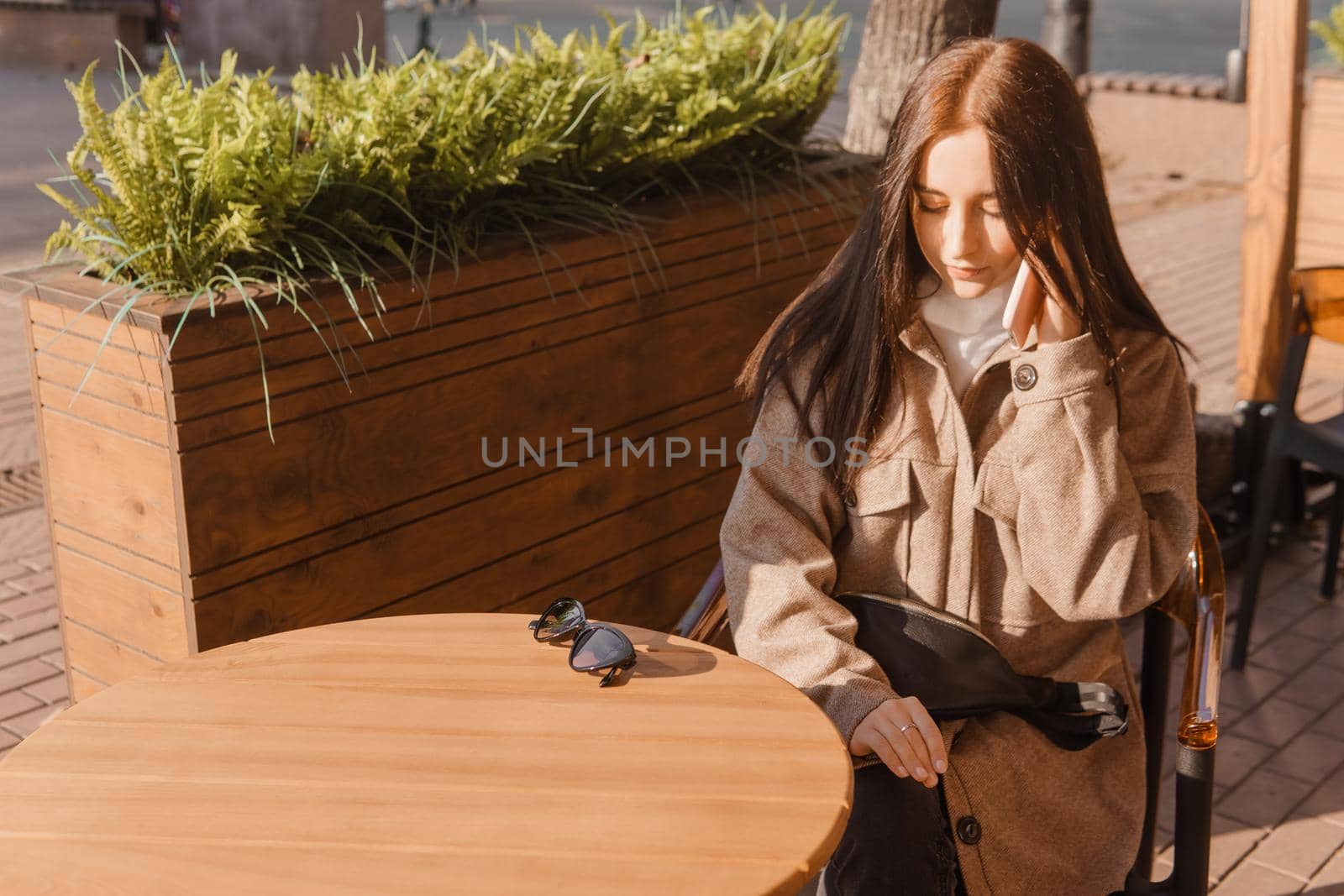 A stylish brunette woman in sunglasses is sitting at a table in an outdoor cafe with coffee and a phone. The girl is talking on the phone. Autumn walk.