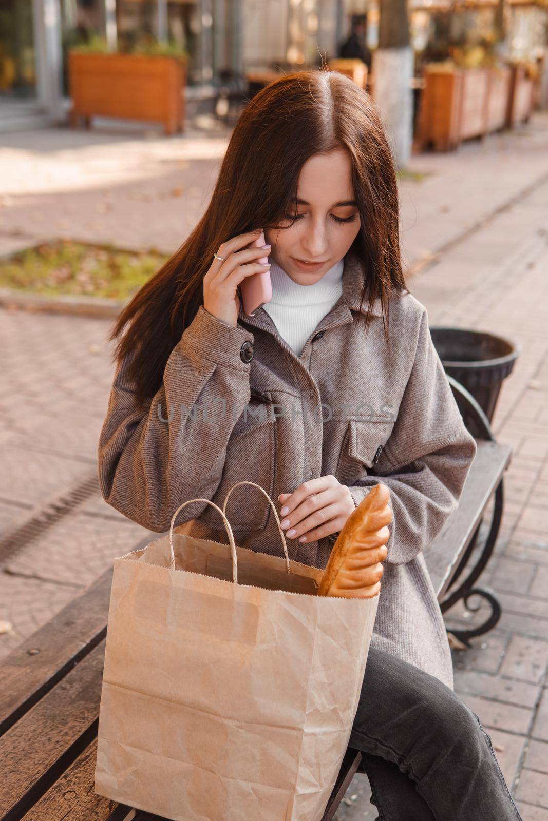 A stylish brunette woman walks around the autumn city. The brunette is sitting on a bench outside with a glass of coffee and a phone, holding a craft bag with a baguette.