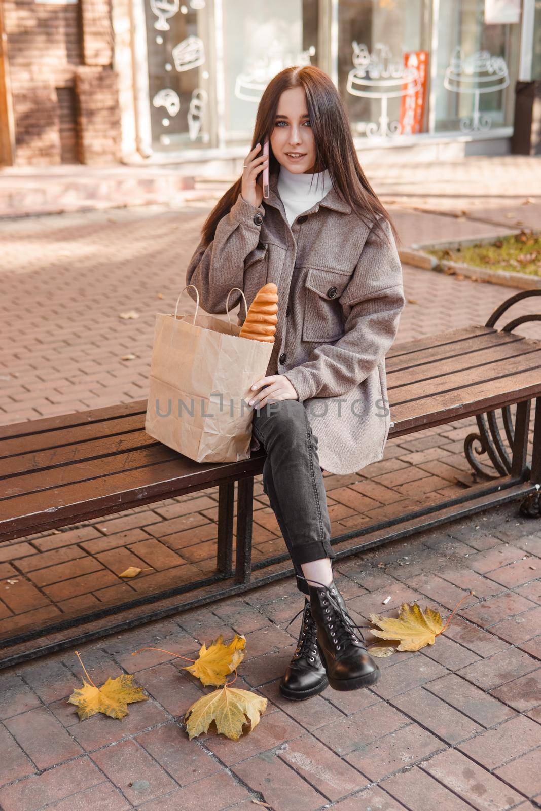 A stylish brunette woman walks around the autumn city. The brunette is sitting on a bench outside with a glass of coffee and a phone, holding a craft bag with a baguette.