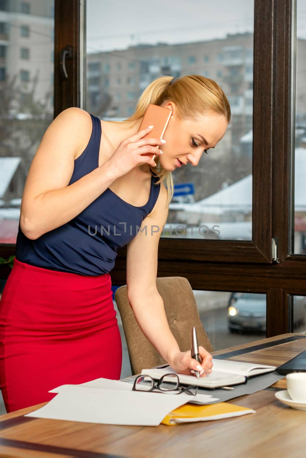 Young businesswoman talks on the phone writing notes standing in office or cafe