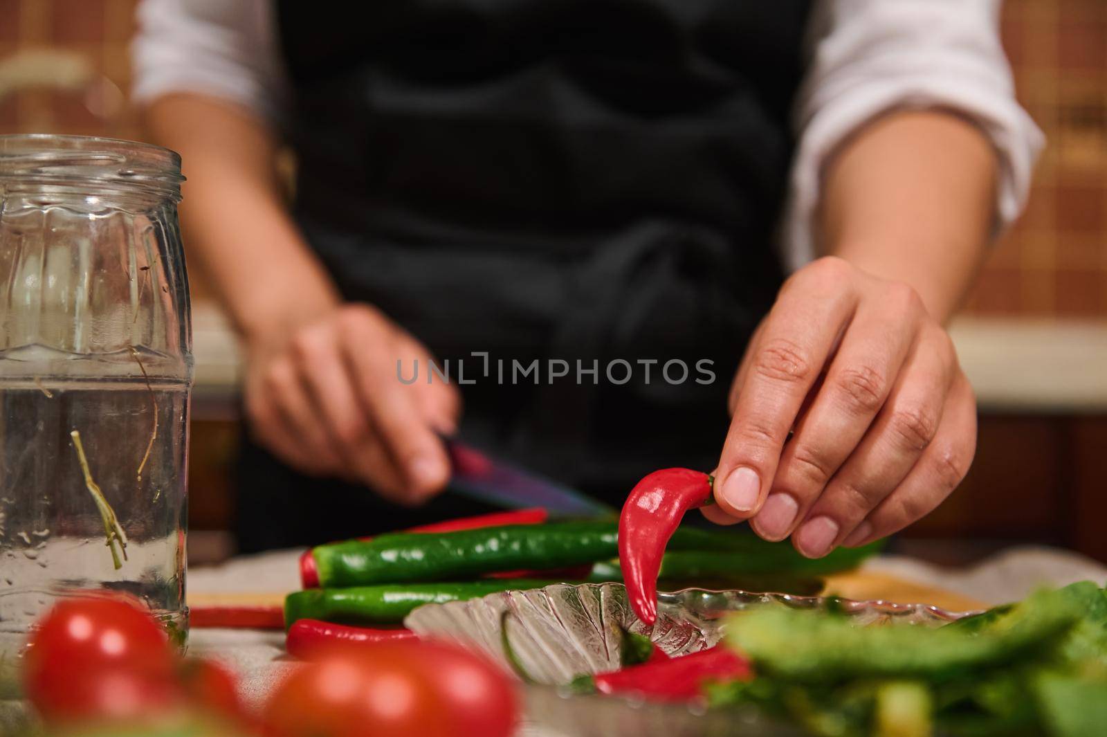 Details: Chef's hand takes red chili peppers out of a plate while slicing raw spicy ingredients during canning food. Raw fresh vegetables are on the table on the blurred foreground. Culinary. Kitchen