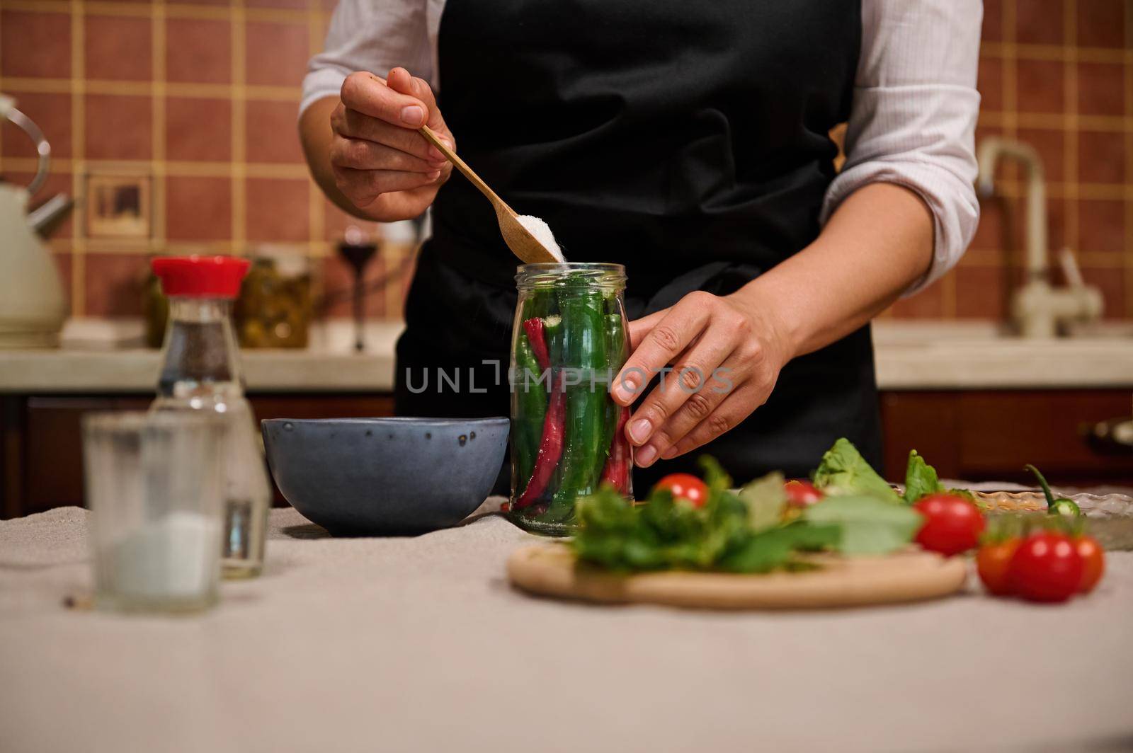 Close-up of a housewife adding a scoop of white sugar in the jar filled with hot chili while preserving for the winter by artgf