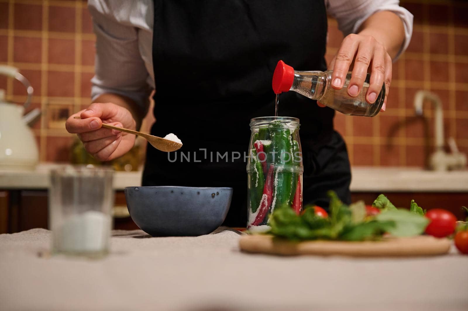 Close-up housewife pouring vinegar into a jar filled with hot chili and fragrant culinary herbs, canning food for winter by artgf