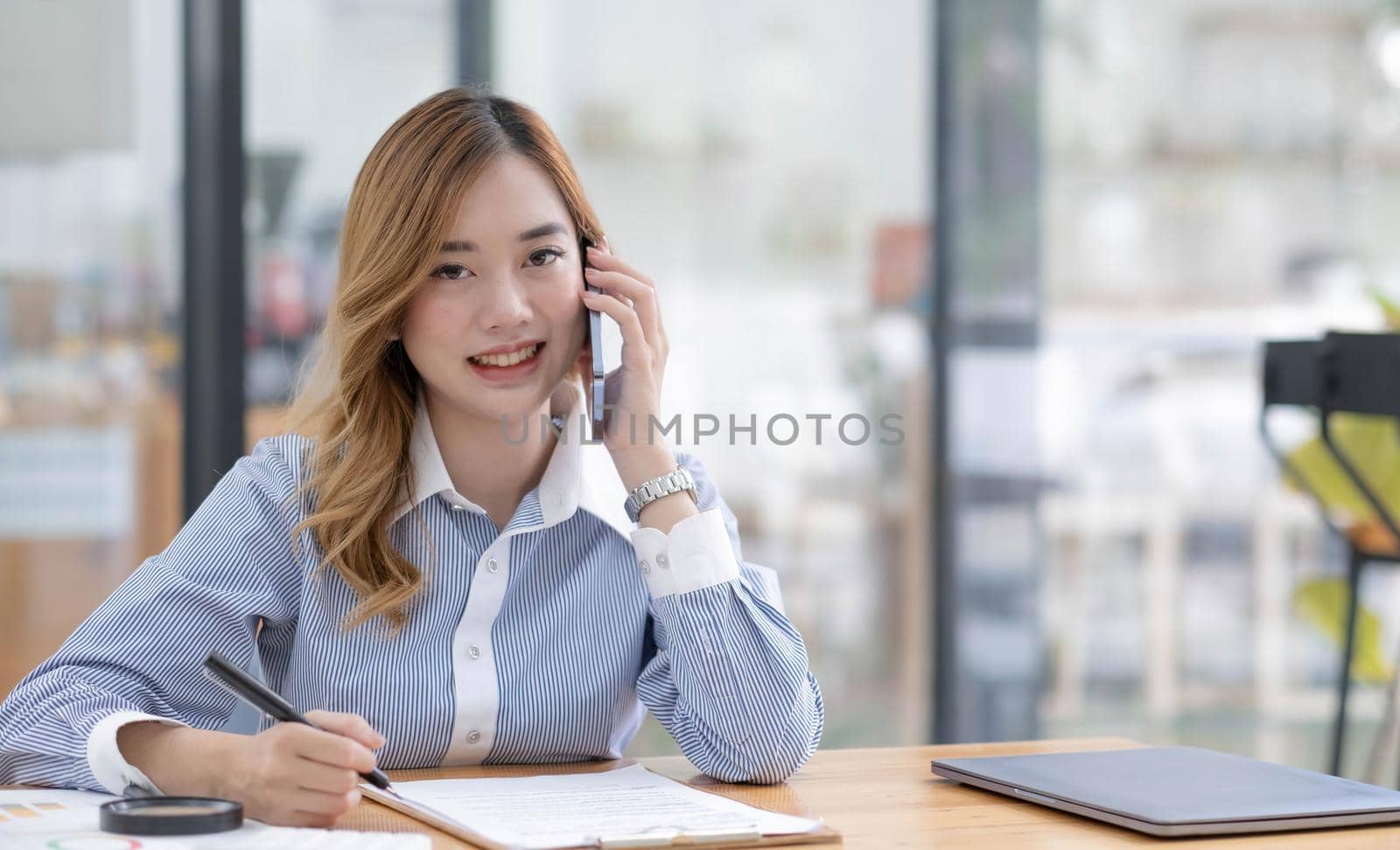 Beautiful Asian businesswoman looking at camera at office by talking on the phone while working at the office by wichayada