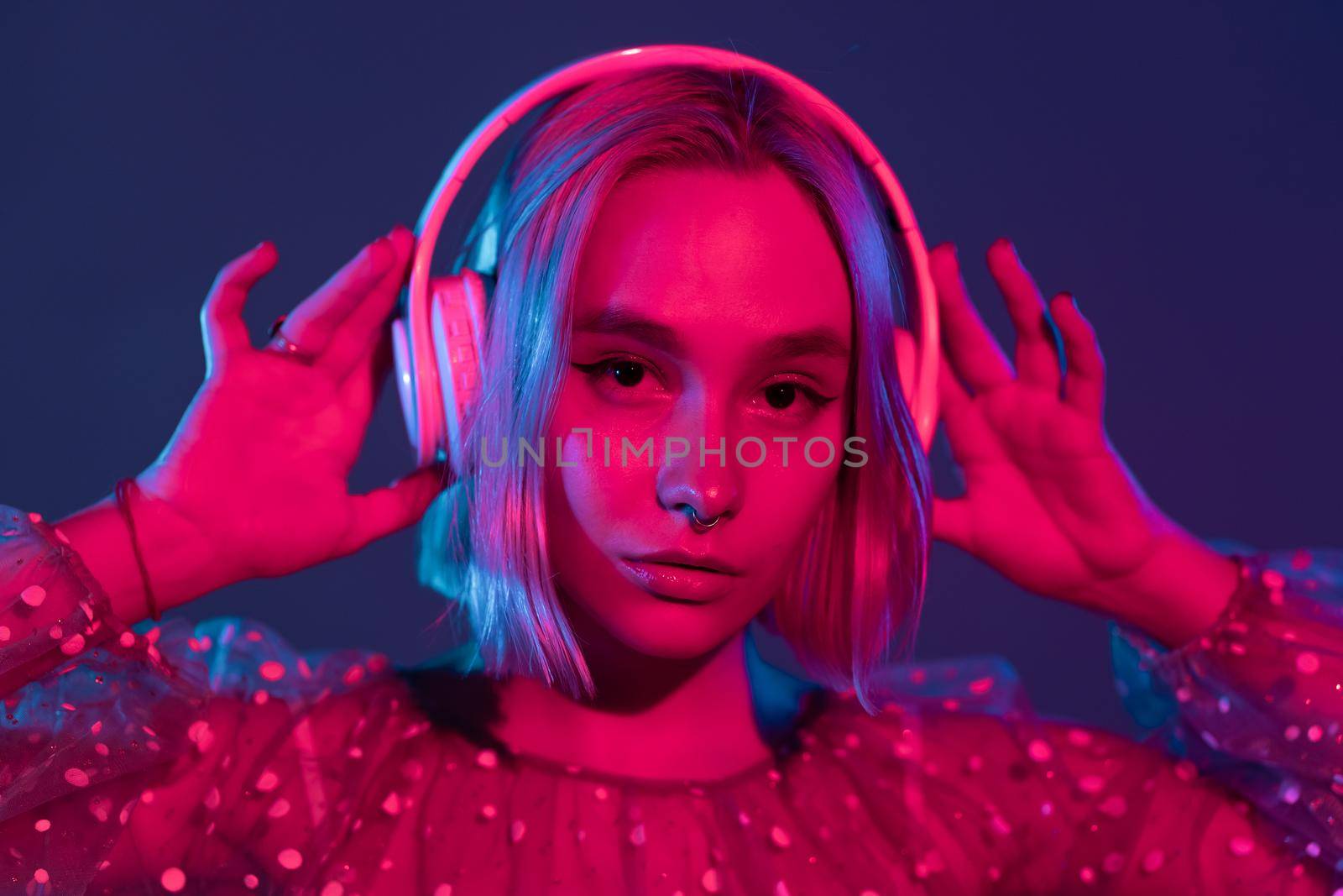 Gorgeous lady with dyed hair listening music in headphones and singing with neon light background. Charming hipster girl dancing with eyes closed