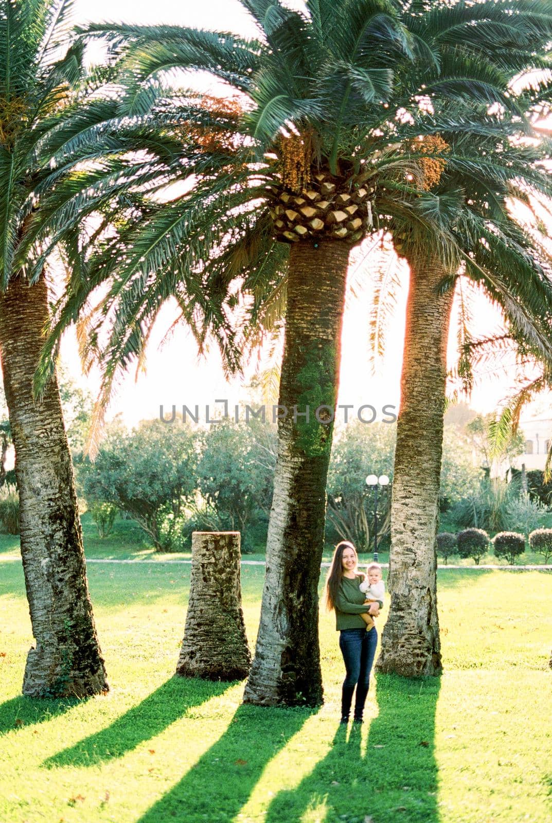 Mom with a baby in her arms stands under huge palm trees on a green lawn by Nadtochiy