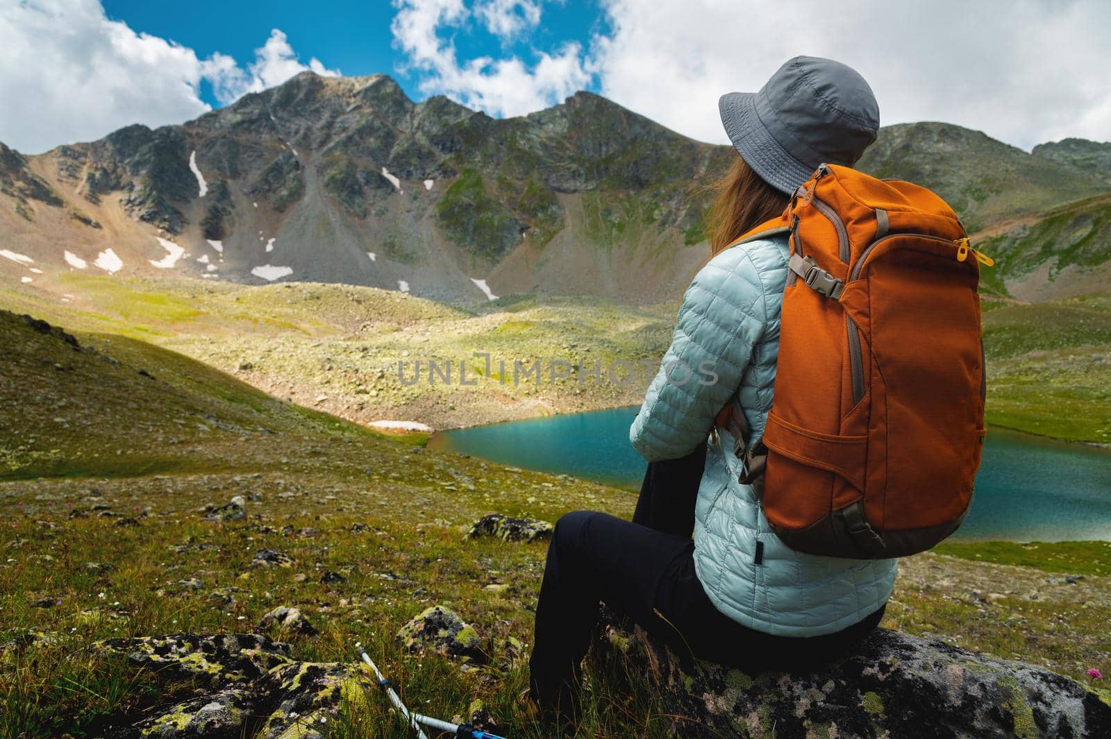 A beautiful young woman sits with her back to the camera on a stone by the lake against the backdrop of high mountains, with a backpack after trekking.