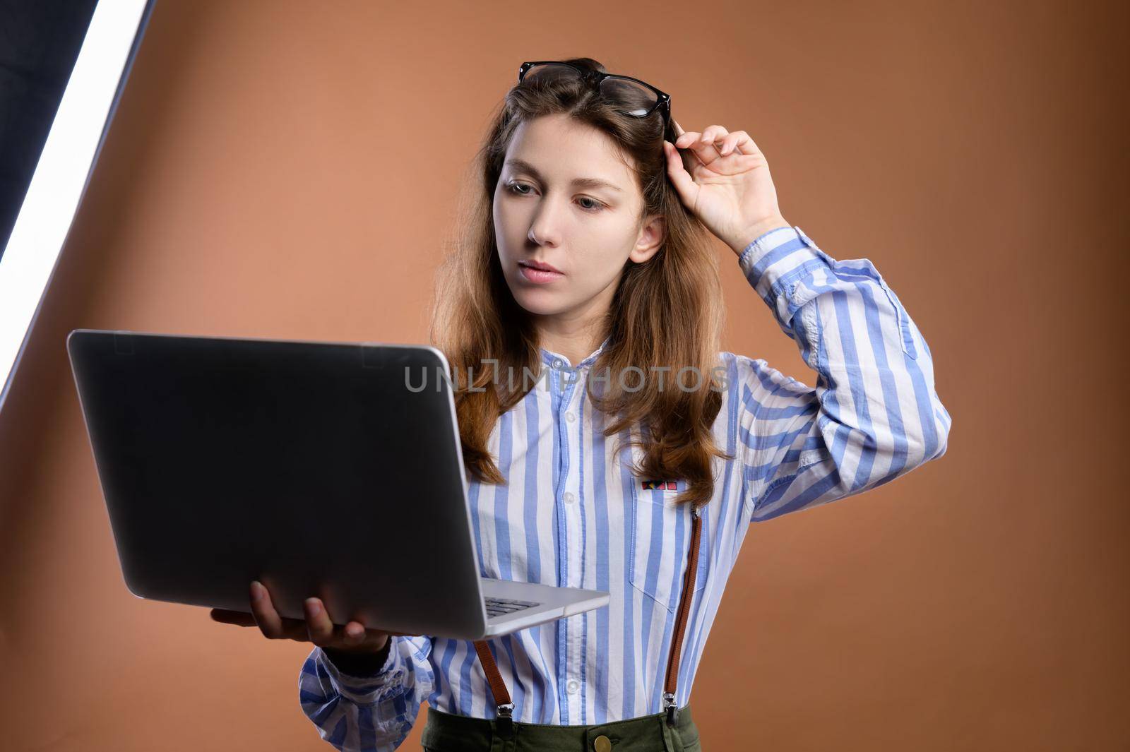 Portrait of a pensive beautiful stylish brunette young woman in glasses, standing, watching and reading the news on the screen of her laptop. shooting indoors, studio.