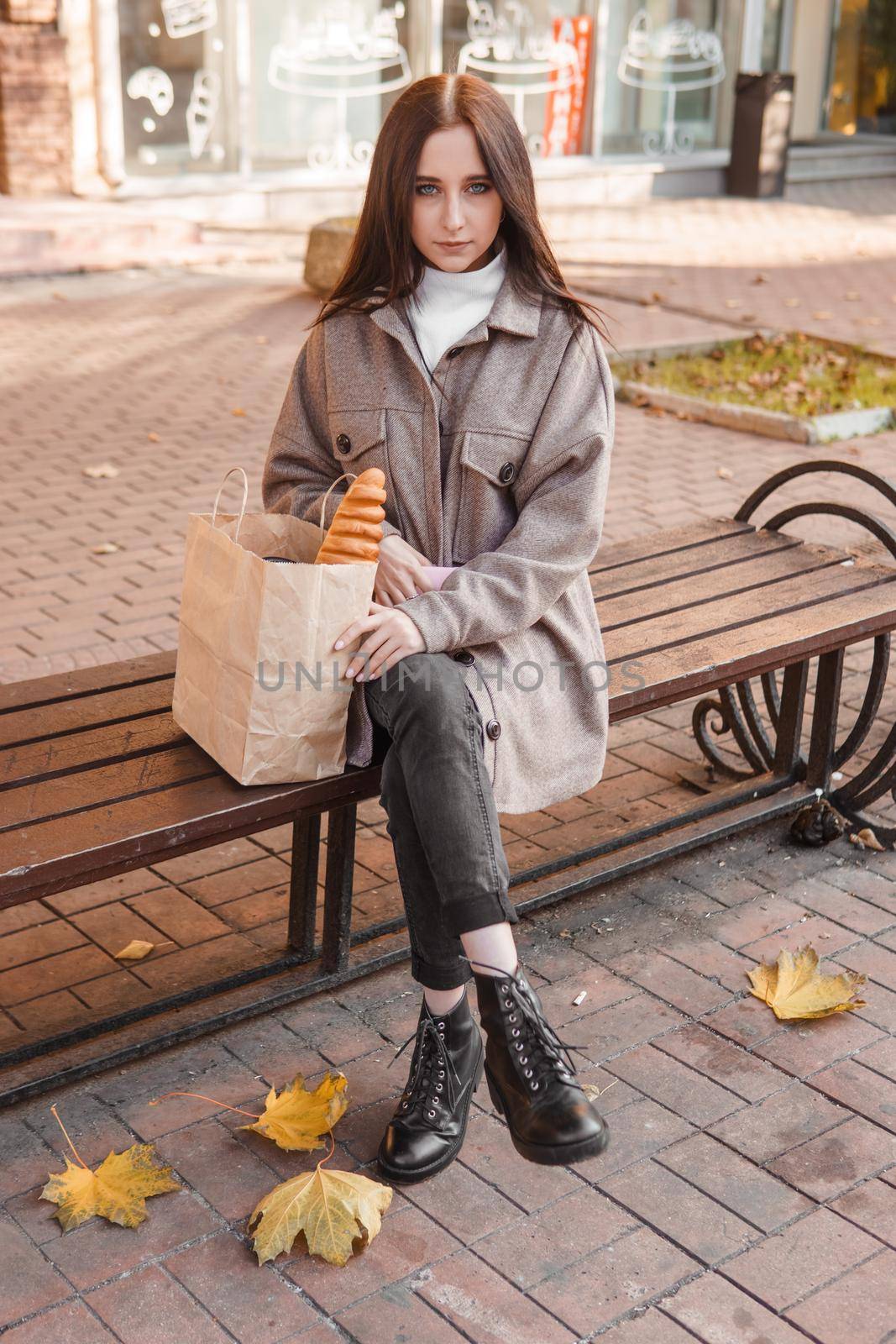 A stylish brunette woman walks around the autumn city. The brunette is sitting on a bench outside with a glass of coffee and a phone, holding a craft bag with a baguette.