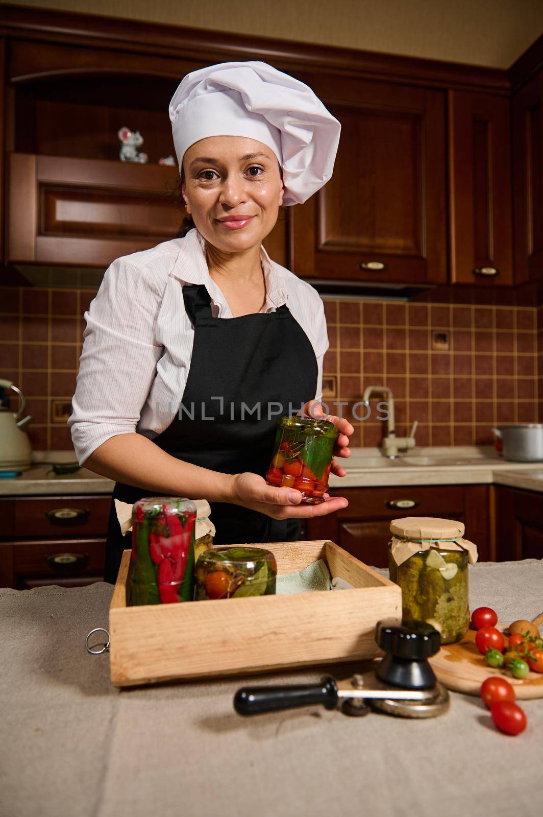 Pretty housewife holds a jar of fermented cherry tomatoes upside down, smiling at camera while standing at kitchen table by artgf