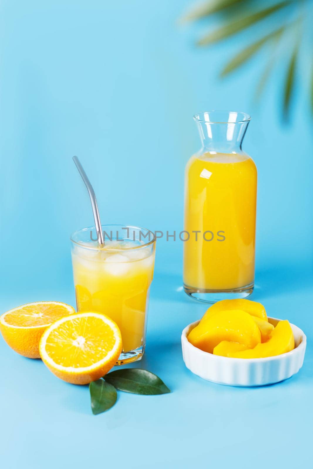 A glass with orange juice, orange and mango slices on a blue background.