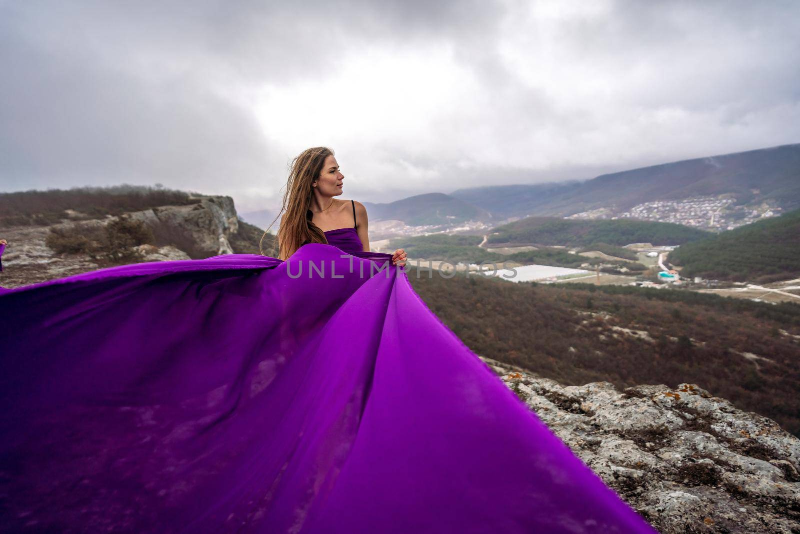 A woman with long hair is standing in a purple flowing dress with a flowing fabric. On the mountain against the background of the sky with clouds. by Matiunina