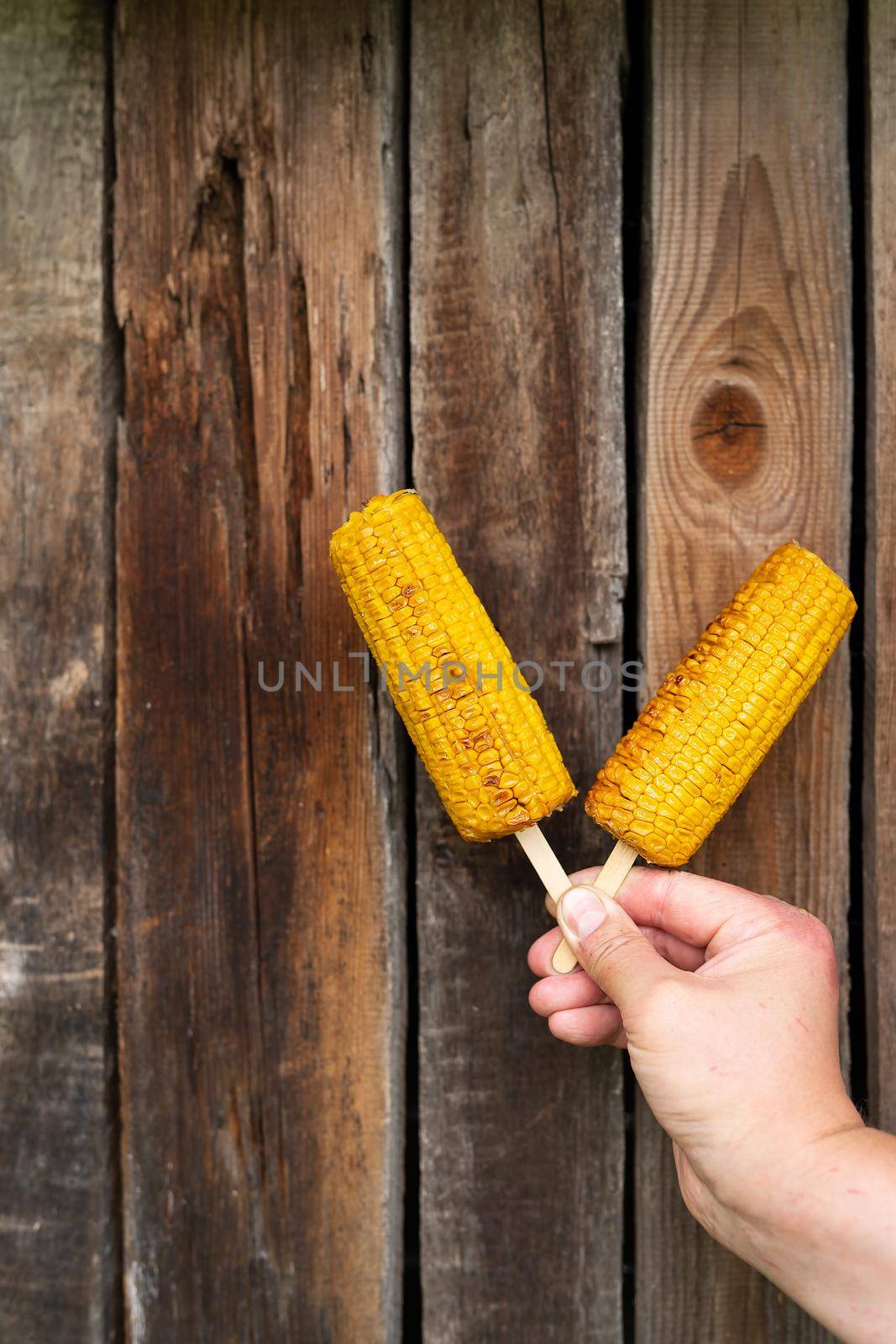 In his hands, a man holds fried sweet corn with salt and butter on a stick on a grill against the background of a wooden old wall. Summer vegan dinner or snack, grill cooking. Vertical photo. by sfinks