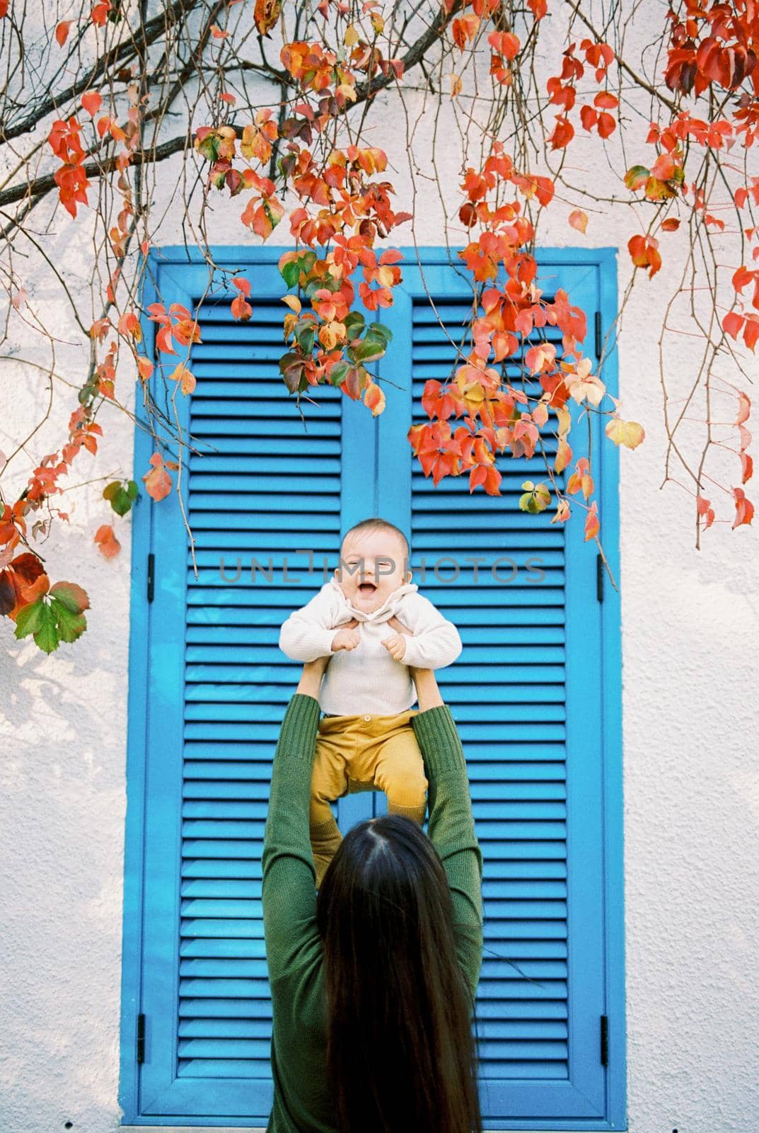 Mom holds a laughing baby high above her head near the blinds on the window. High quality photo