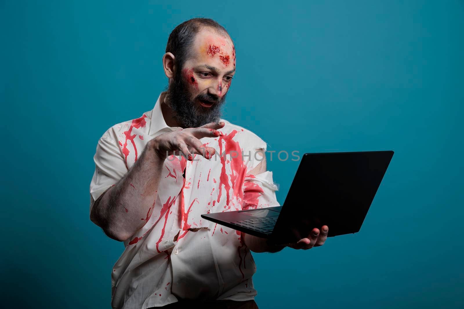 Halloween zombie using laptop computer in studio, looking creepy and dangerous browsing internet while being spooky and apocalyptic. Brain eating corpse with bloody scars using social media on pc.