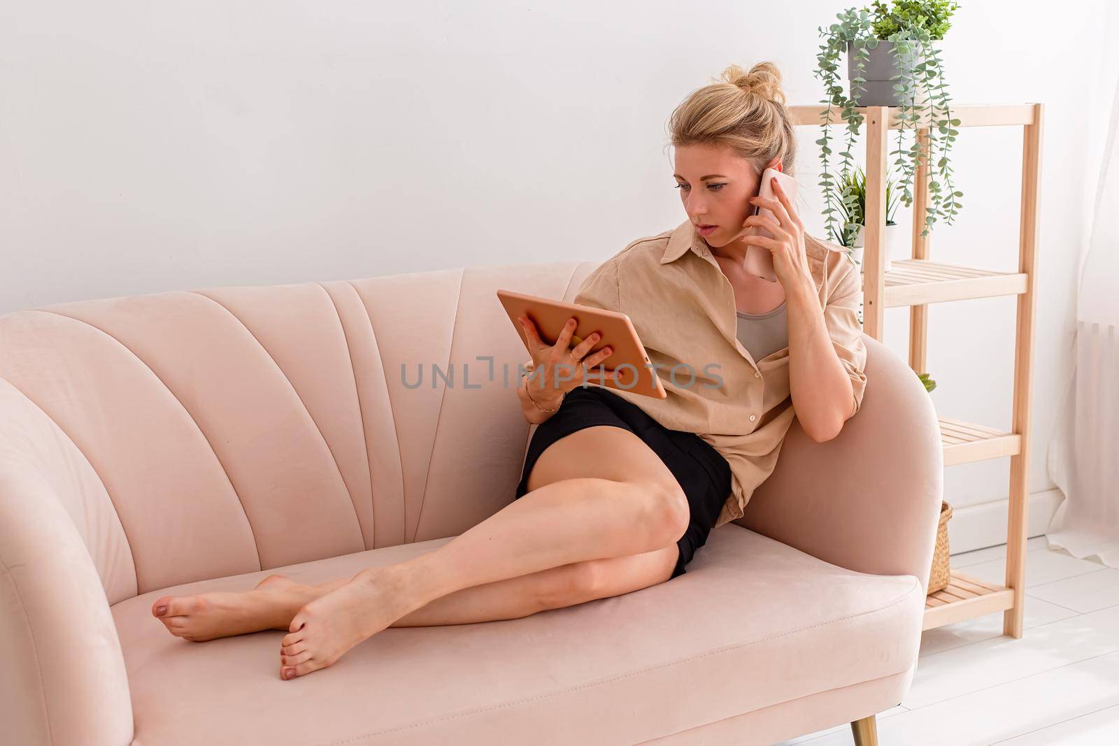 Blonde woman, 30-40 years old, looking at a digital tablet and talking on a mobile phone while lying comfortably on a beige sofa in a bright living room.