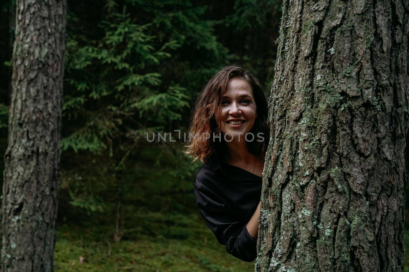 A young woman in a coniferous forest looks out from behind a tree. Happy young woman looking at the camera and smiling