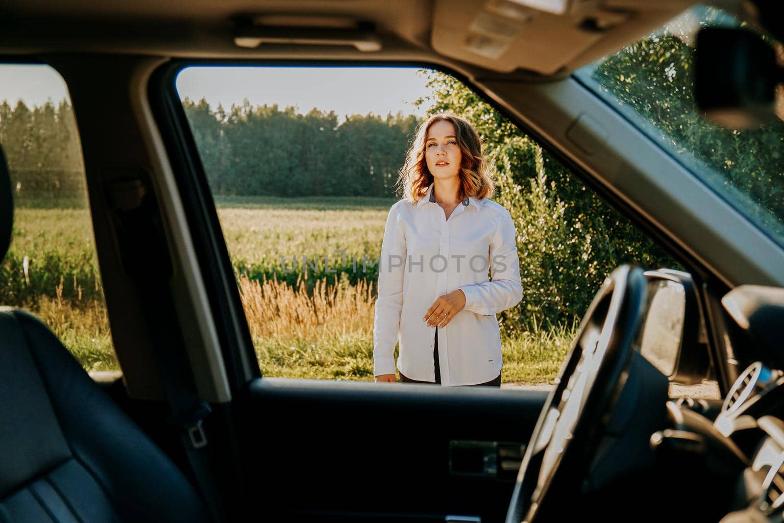 A beautiful young woman in a white shirt is resting outside the city. Outdoors near the forest and field. Photo through the car window. Car trip