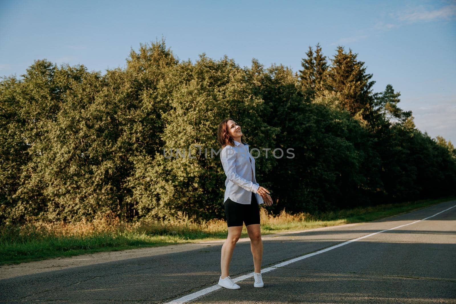 A red-haired woman in a white shirt walks along the road among the forest by natali_brill
