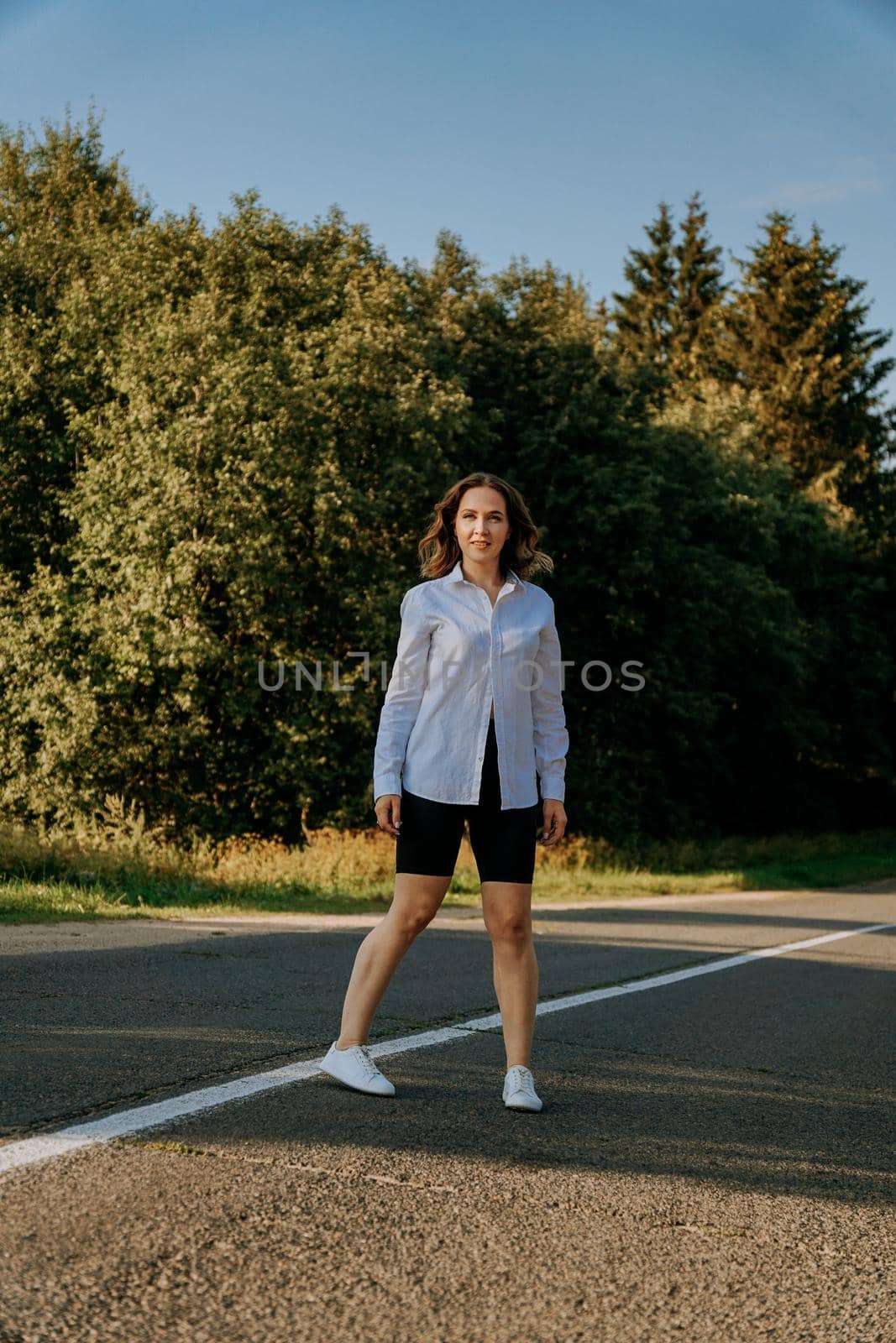 A red-haired woman in a white shirt walks along the road among the forest on a summer sunny day. A trip out of town.