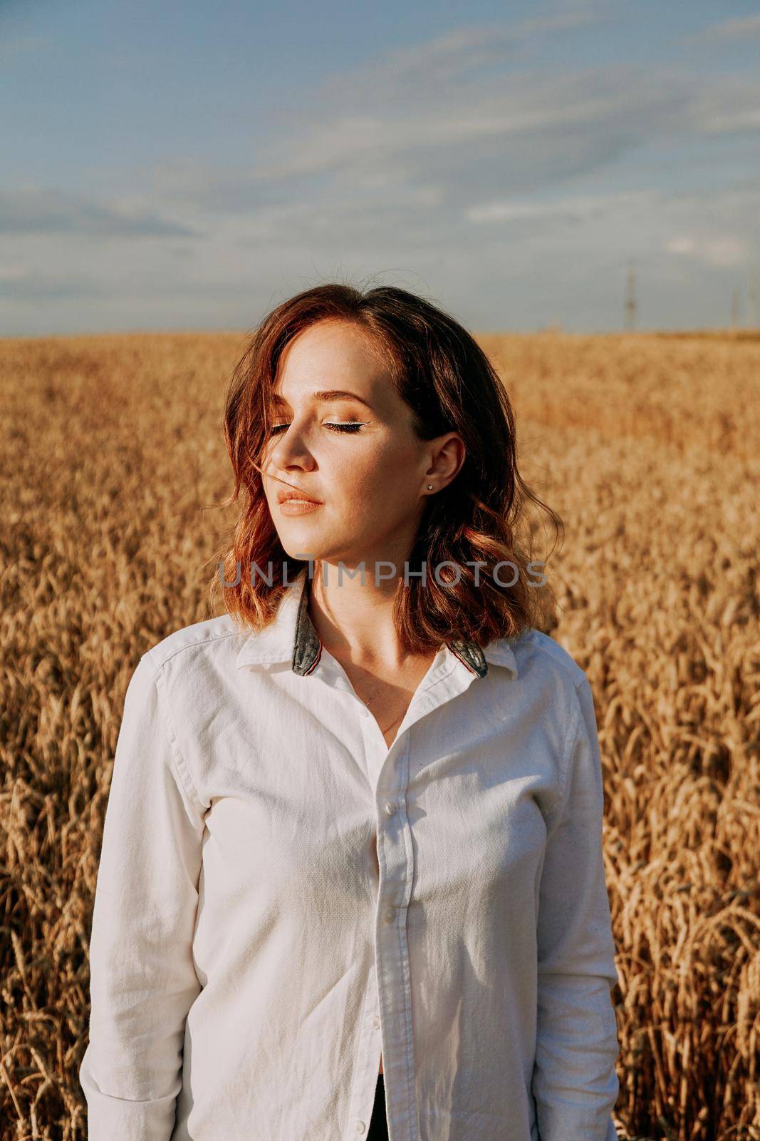 Portrait of a beautiful red-haired girl in a white shirt. She stands in a rye field on a sunny day. Calm facial expression. The concept of appeasement. Vertical photo