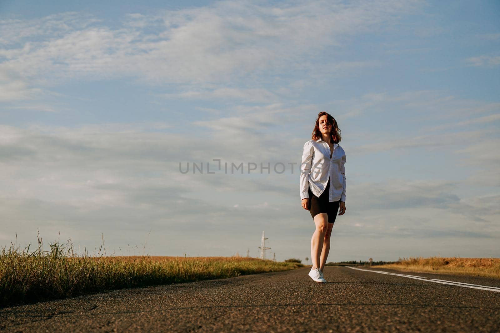 A red-haired woman in a white shirt walks along the road among the fields on a summer sunny day. A trip out of town