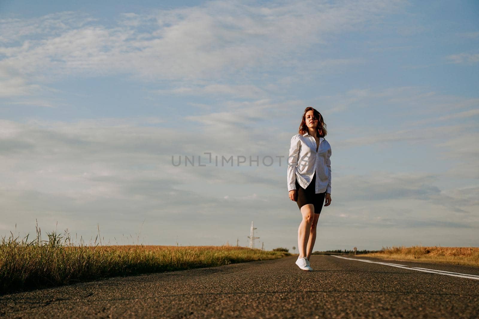 A red-haired woman in a white shirt walks along the road among the fields by natali_brill