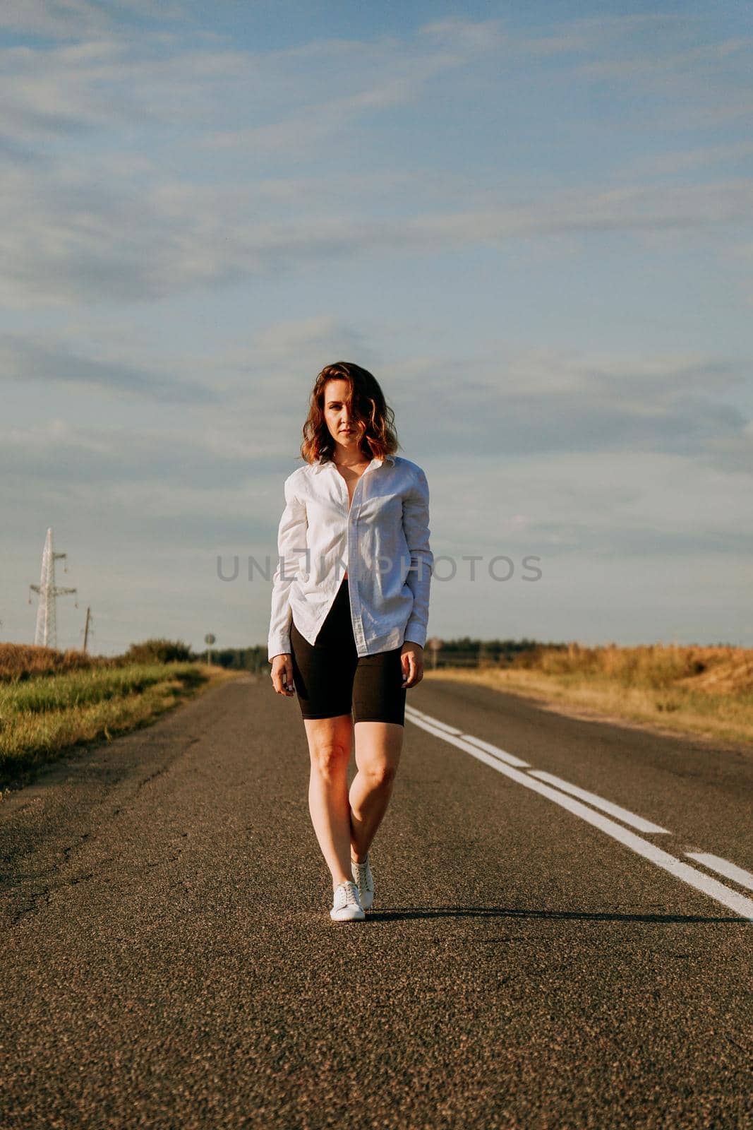 A red-haired woman in a white shirt walks along the road among the fields by natali_brill