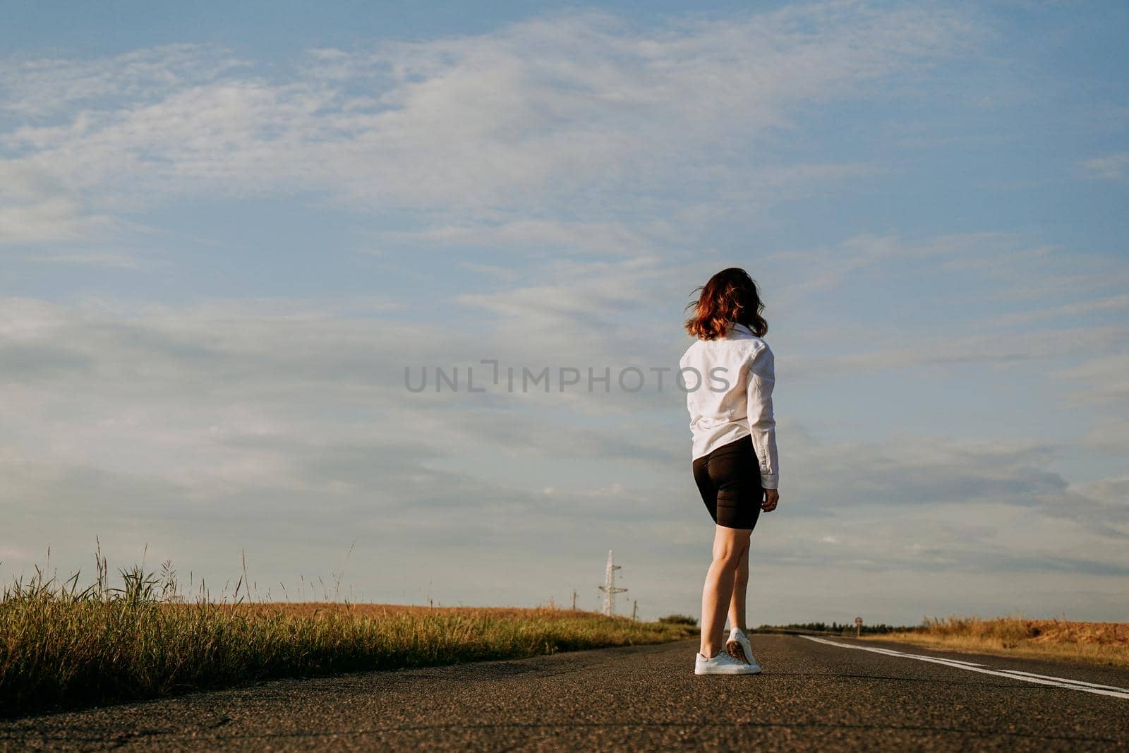 A red-haired woman in a white shirt walks along the road among the fields by natali_brill