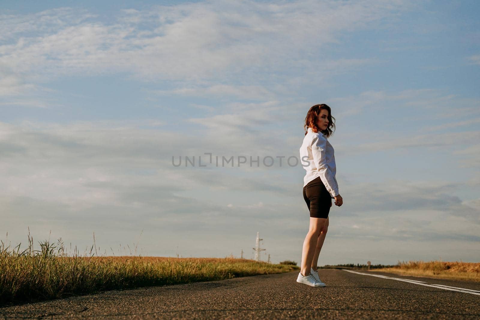 A red-haired woman in a white shirt walks along the road among the fields on a summer sunny day. A trip out of town