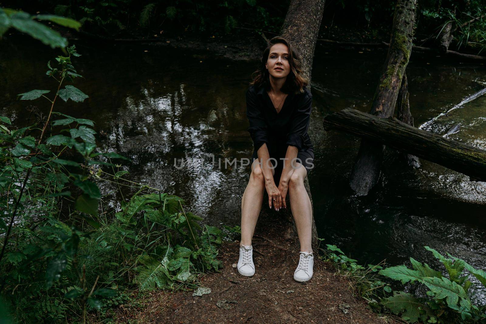 Woman in black clothes. Walk in a dark coniferous forest. Tracking and trip. Girl posing next to a tree above water