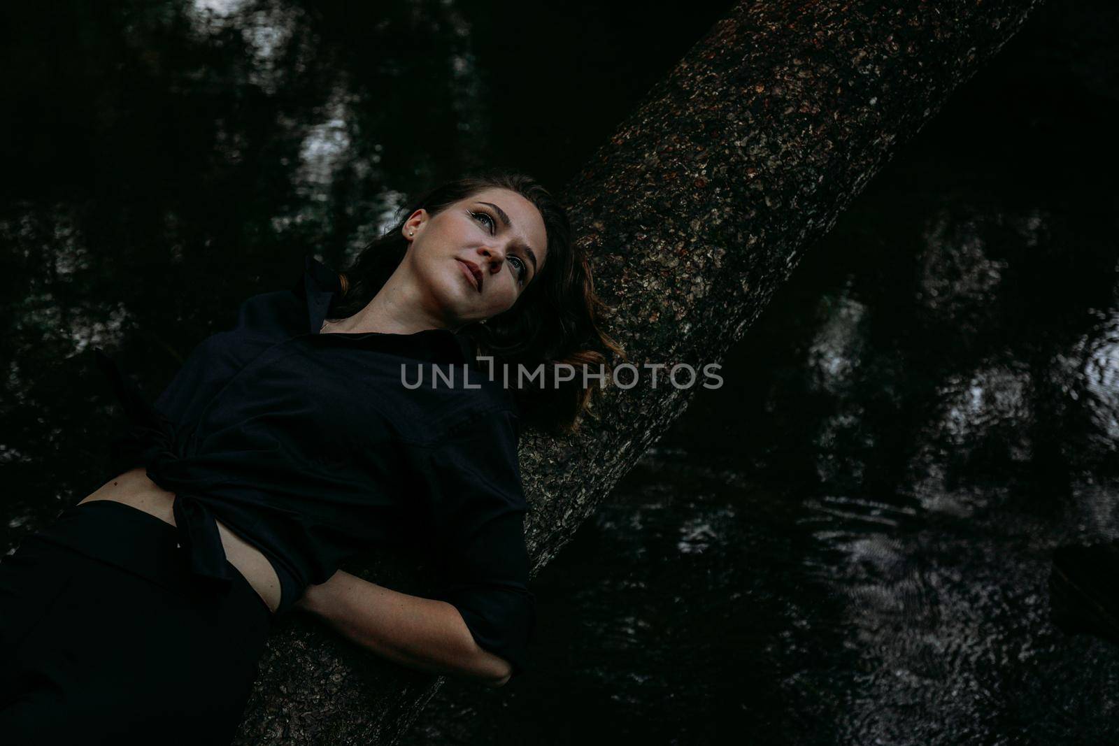 Woman in black clothes on a background of water, lies on a tree. Photo in the forest, the atmosphere of sadness and depression