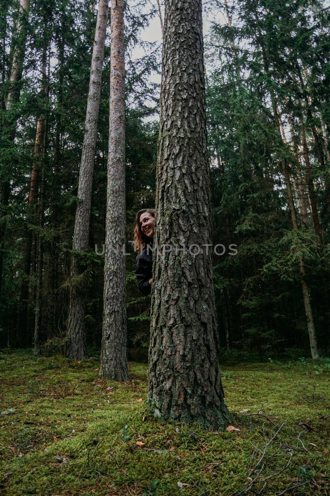 A young woman in a coniferous forest looks out from behind a tree by natali_brill