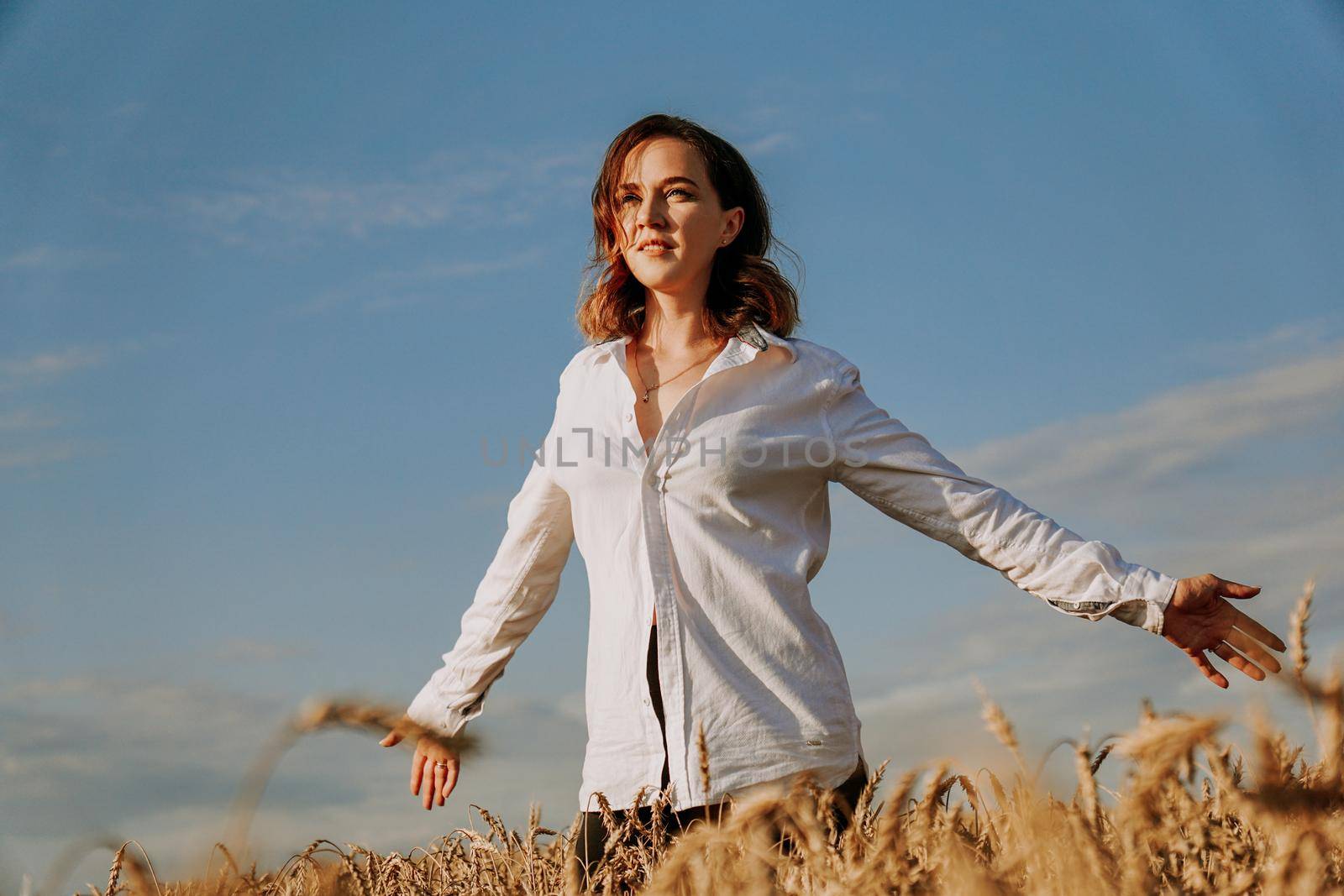 Happy young woman in a white shirt in a wheat field. Sunny day. Girl smiling, happiness concept. Hands to the side