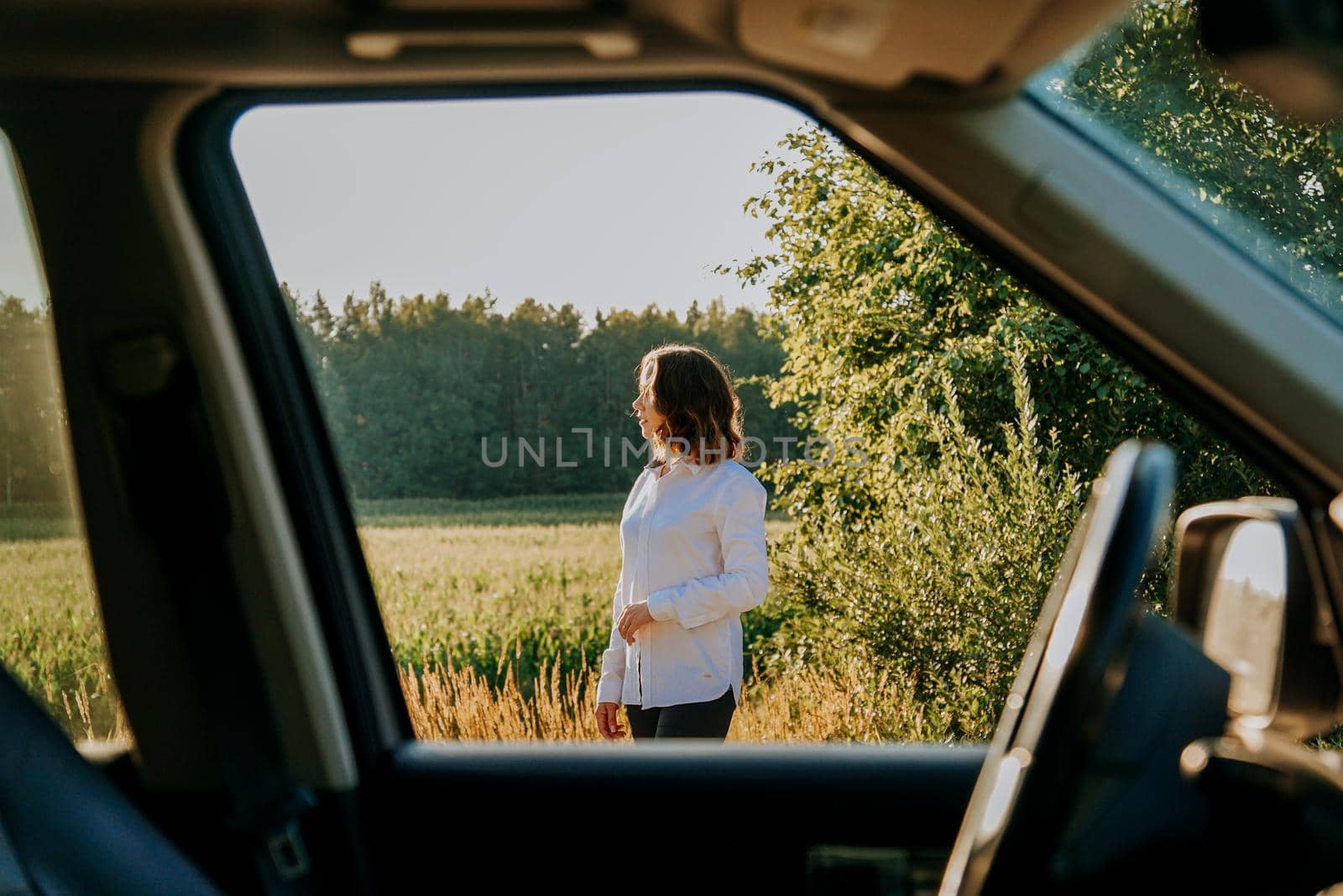 A beautiful young woman in a white shirt is resting outside the city. Outdoors near the forest and field. Photo through the car window. Car trip