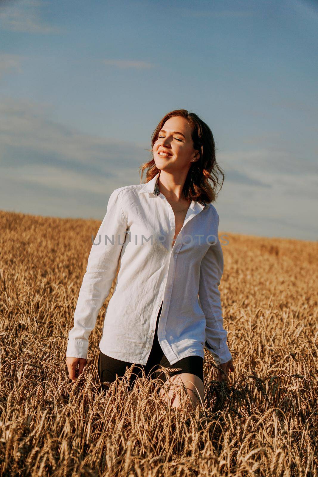 Portrait of a beautiful red-haired girl in a white shirt. She stands in a rye field on a sunny day. Calm facial expression. The concept of appeasement