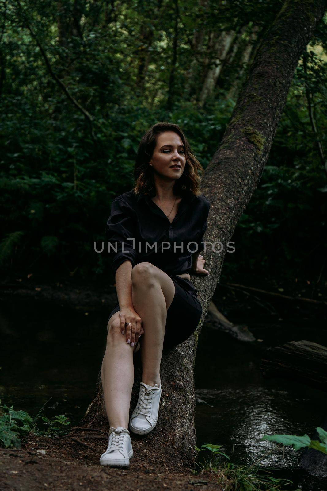 Woman in black clothes. Walk in a dark coniferous forest. Tracking and trip. Girl posing next to a tree above water