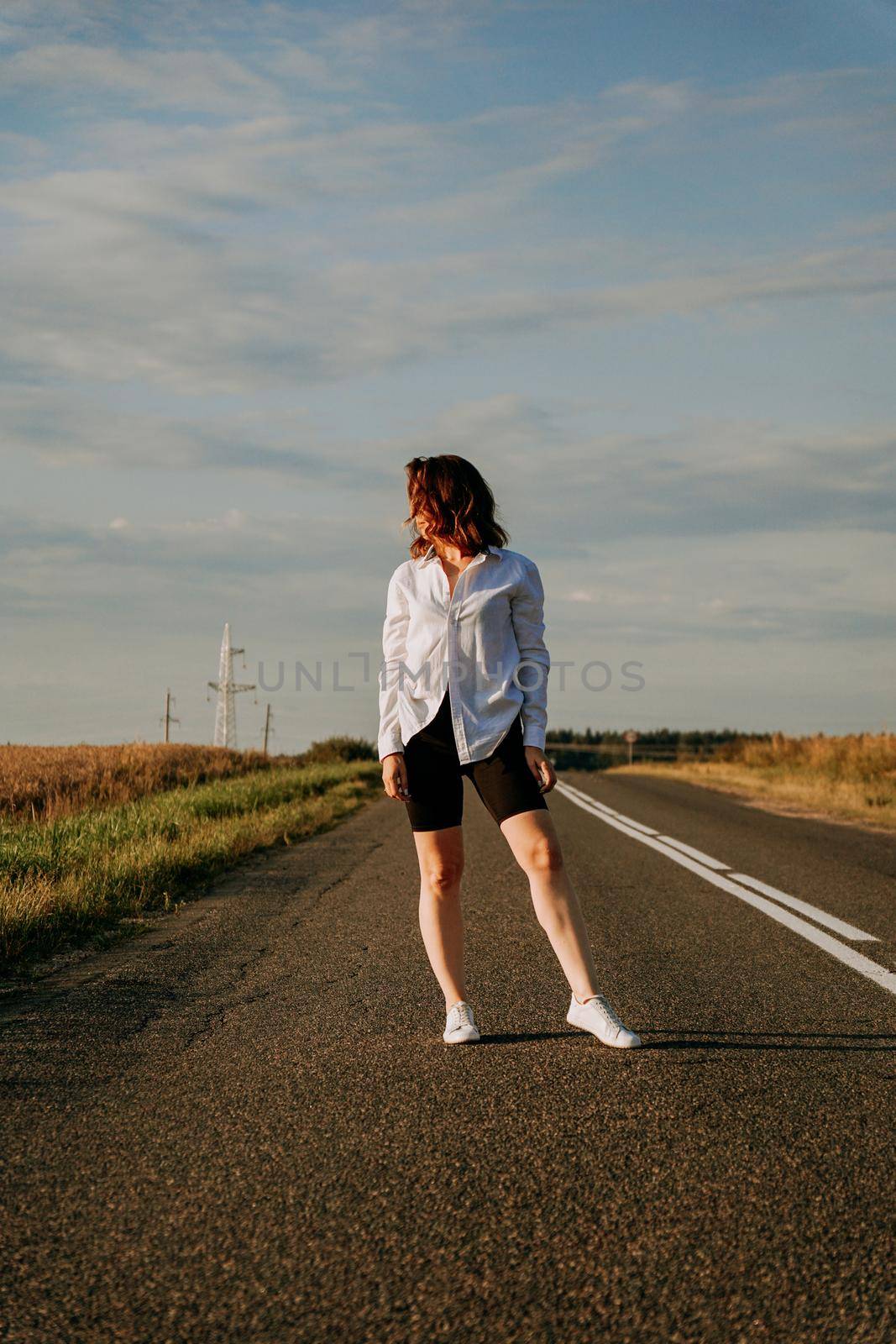 A red-haired woman in a white shirt walks along the road among the fields on a summer sunny day. A trip out of town. Vertical photo