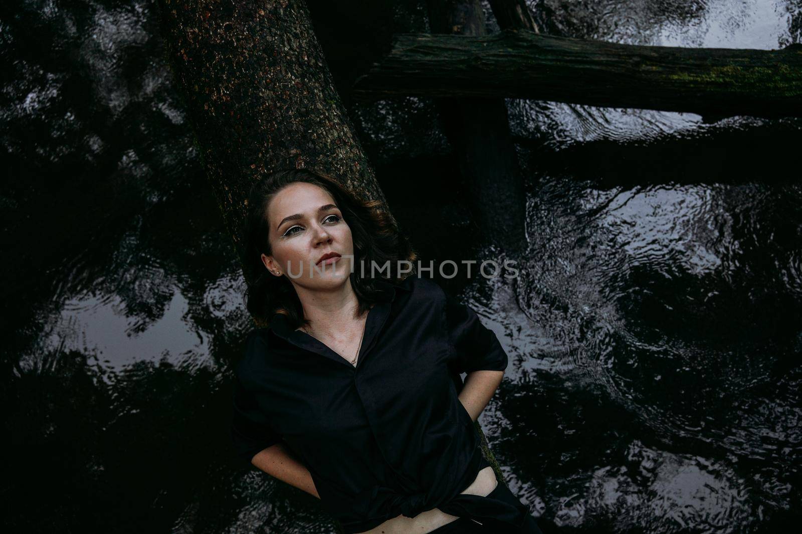 Woman in black clothes on a background of water, lies on a tree. Photo in the forest, the atmosphere of sadness and depression