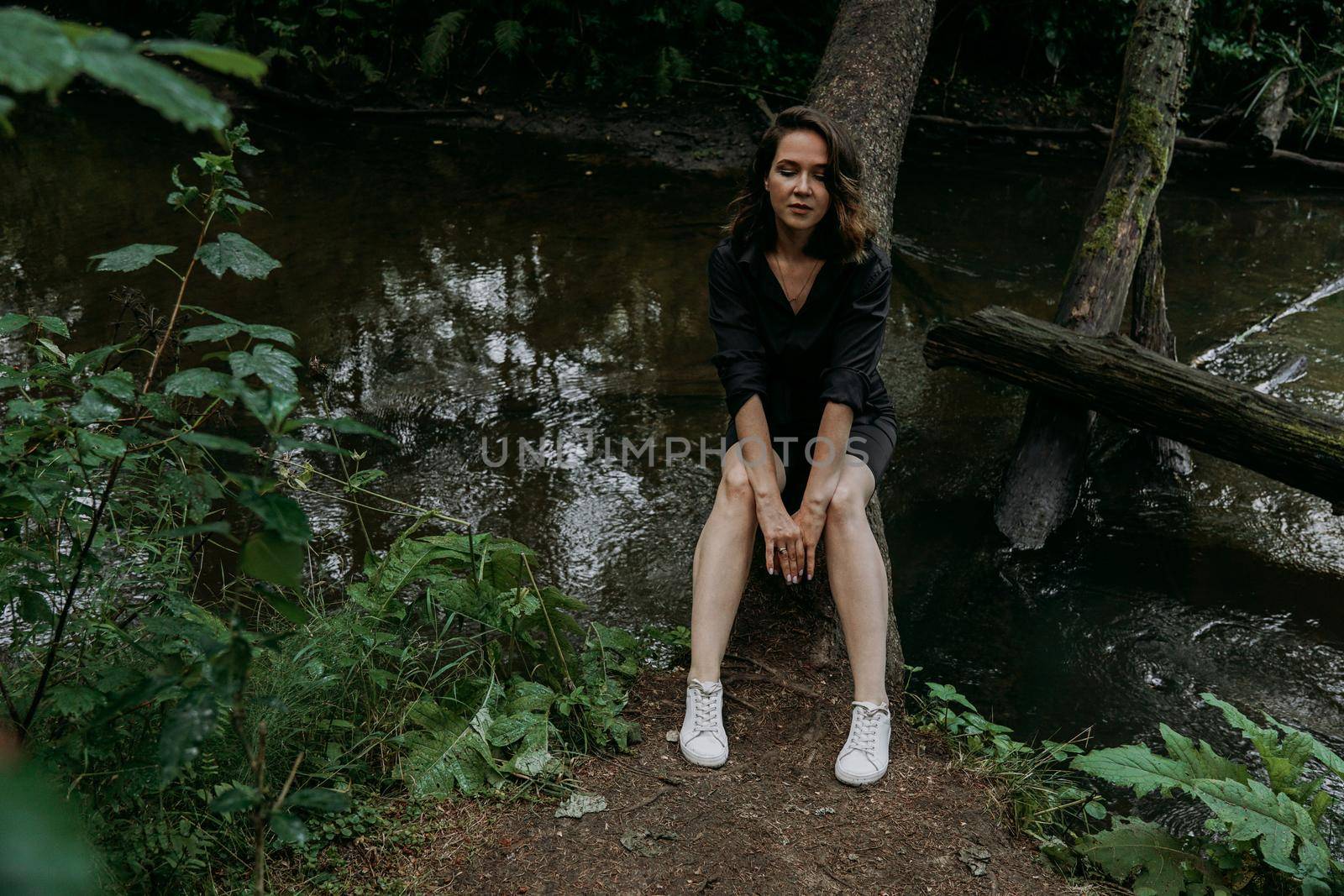 Woman in black clothes. Walk in a dark coniferous forest. Tracking and trip. Girl posing next to a tree above water