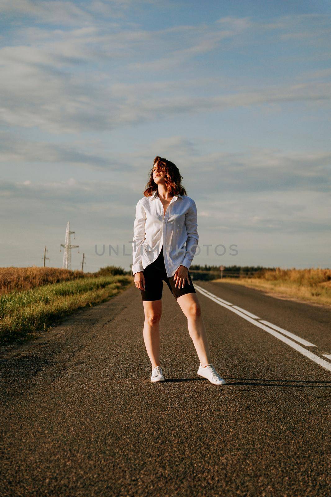 A red-haired woman in a white shirt walks along the road among the fields on a summer sunny day. A trip out of town. Vertical photo