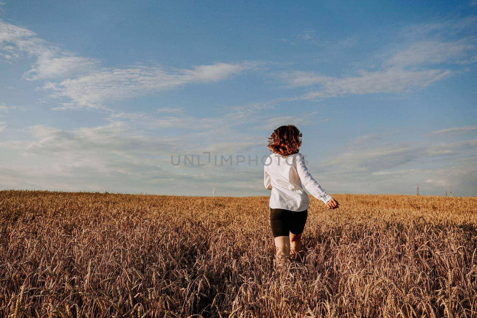 Happy woman in a white shirt runs in a wheat field on a summer day. Happiness and joy concept. Back view