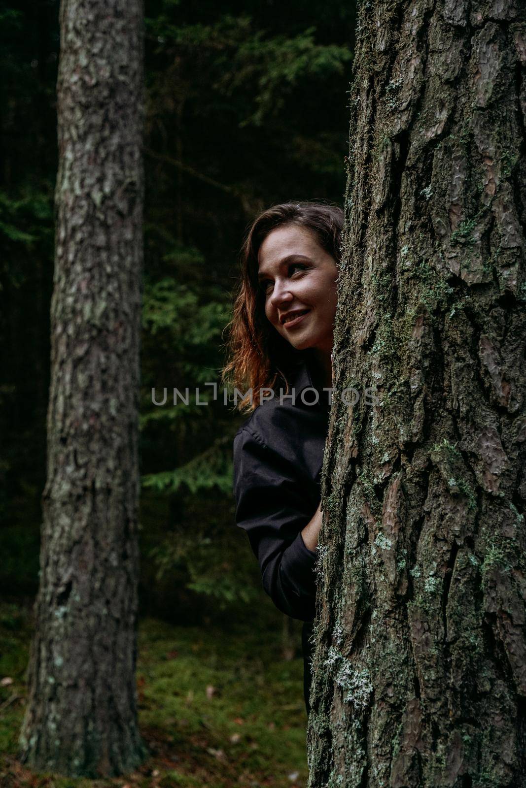 A young woman in a coniferous forest looks out from behind a tree. Happy young woman
