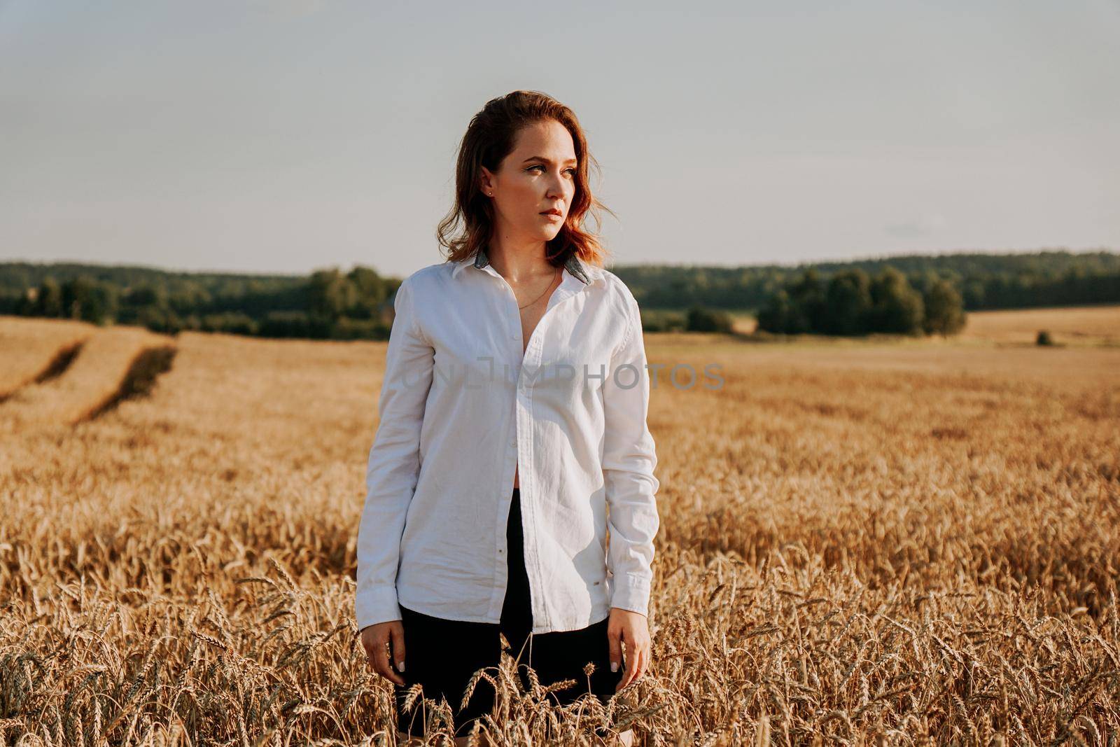 Portrait of a beautiful red-haired girl in a white shirt. She stands in a rye field on a sunny day. Calm facial expression. The concept of appeasement