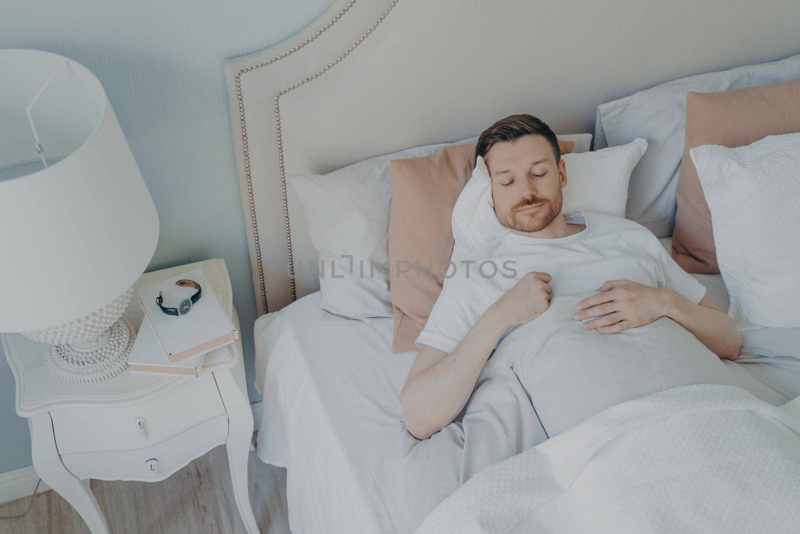Handsome young bearded man peacefully sleeping in spacious comfortable bed in early morning at home, male enjoying sweet dreaming and relaxation during night sleep. View from above