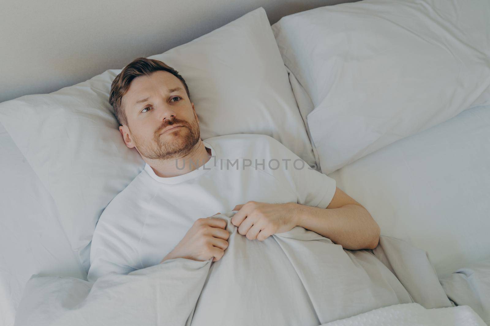 Lonely upset young bearded man lying in bed with open eyes by vkstock