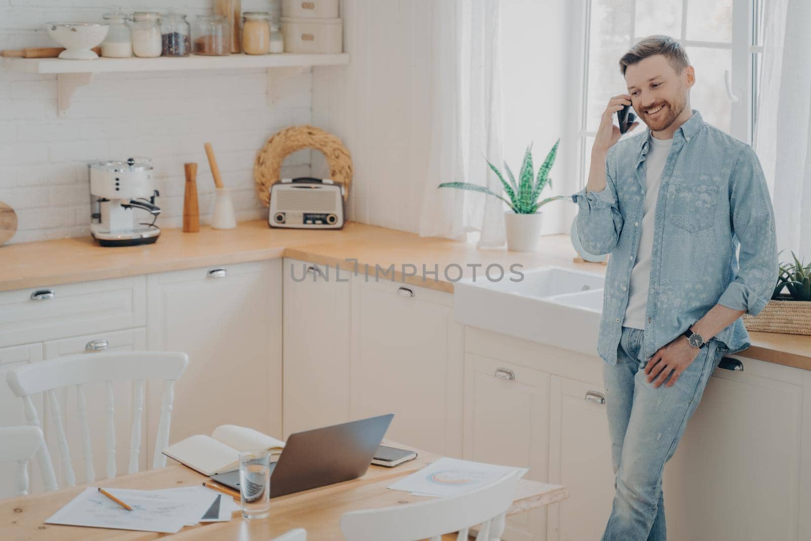 Happy young caucasian man holding cellphone while having conversation with client, standing in modern kitchen and enjoying good business talk while working distantly from home