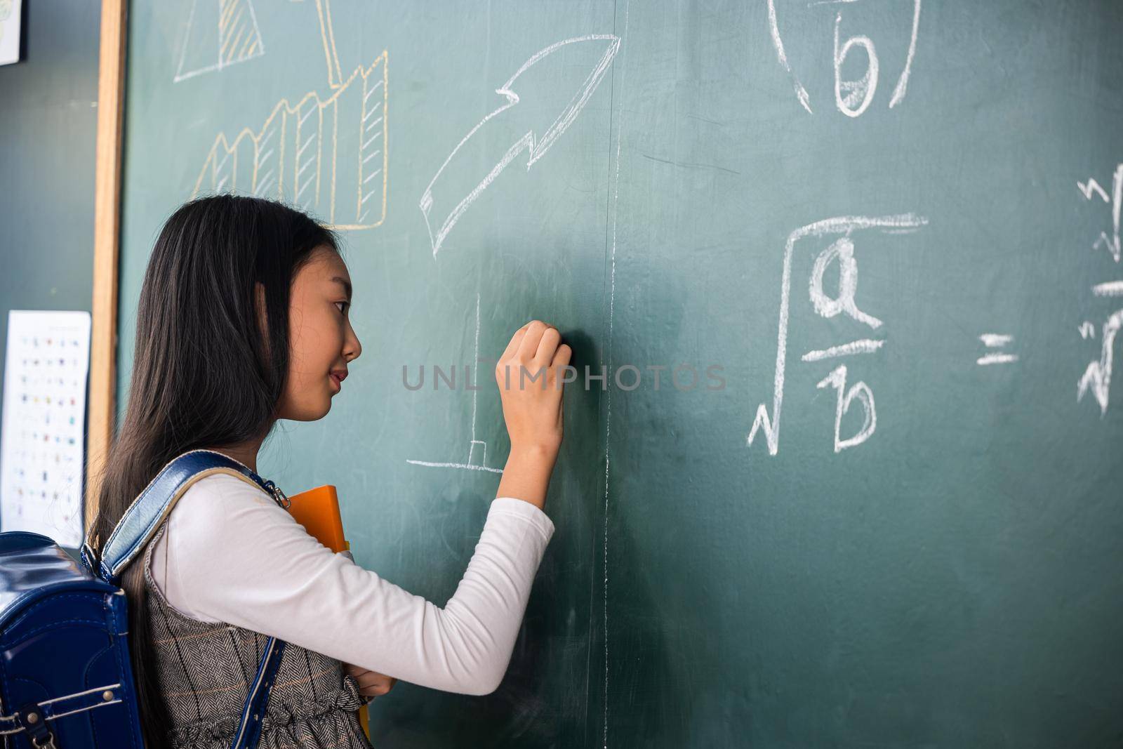 Education. Back view of school girl on science lesson in classroom write an answer on blackboard, primary child is standing in front of class in school writing chalkboard, Back to school concept