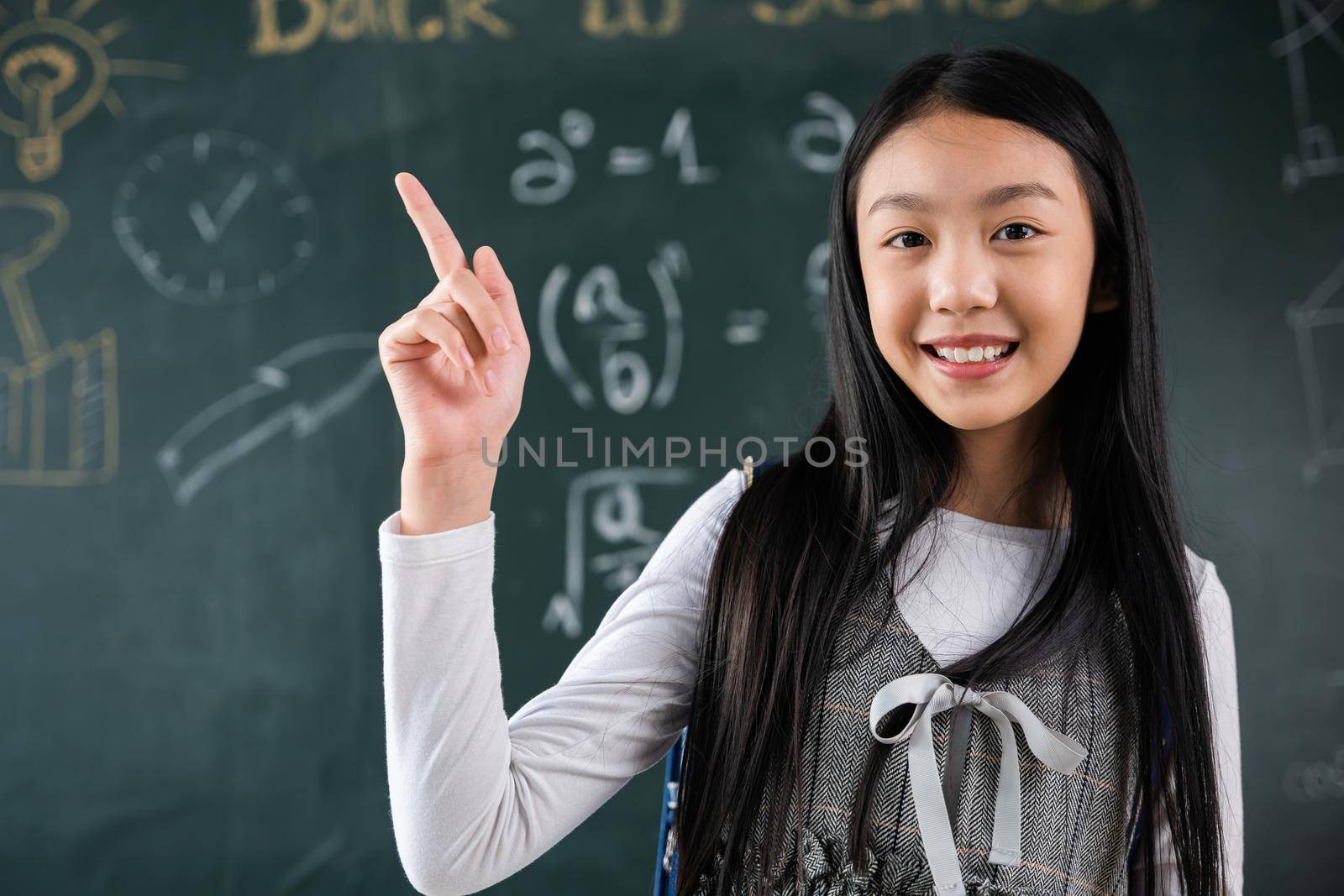Education Back to school concept. Asian school girl in uniform with backpack on classroom pointing up finger on blackboard, primary child is standing front of class in school thinking of great ideas