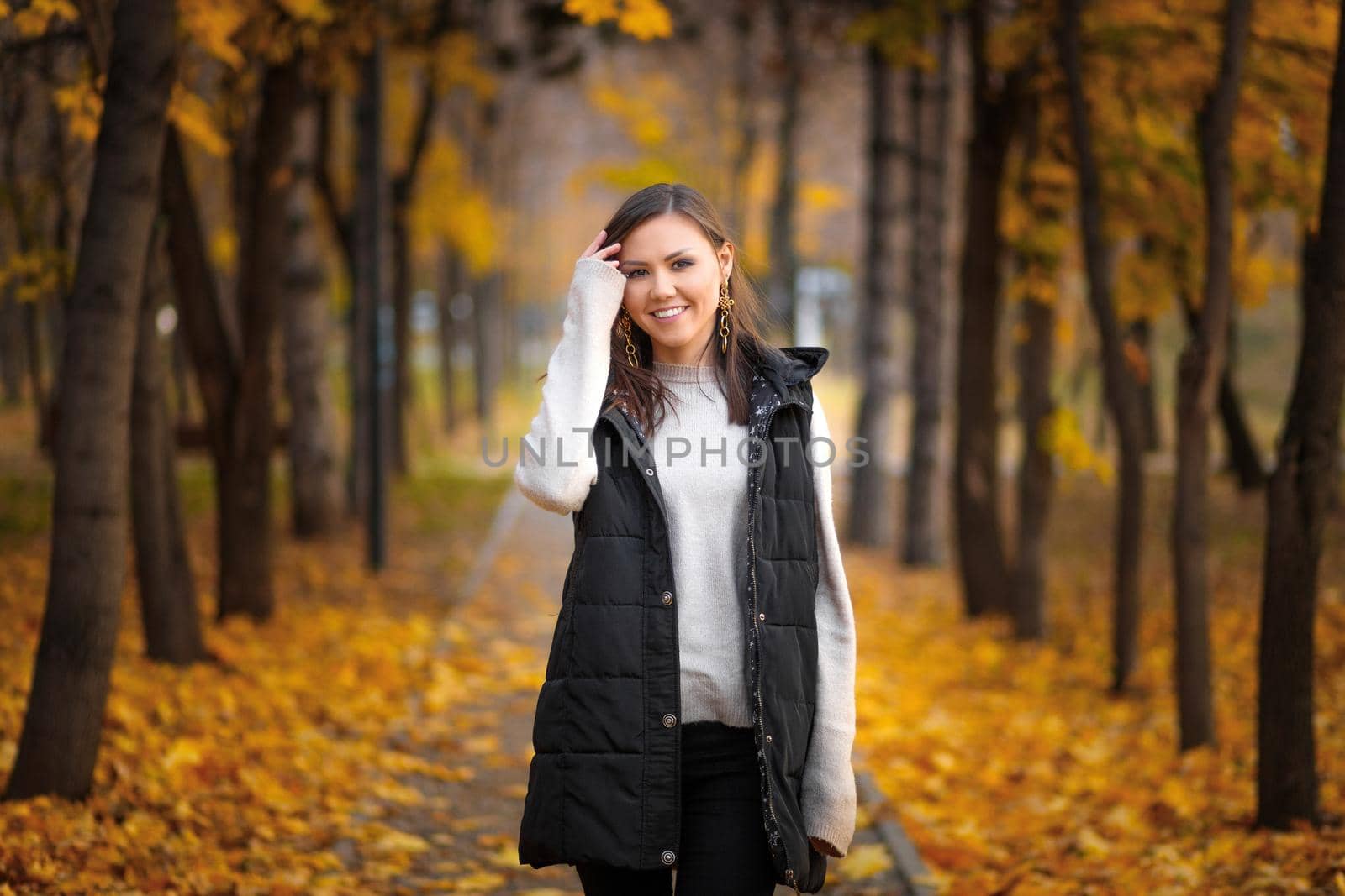 Portrait of a young Central Asian woman in an autumn park on an alley with fallen autumn leaves.